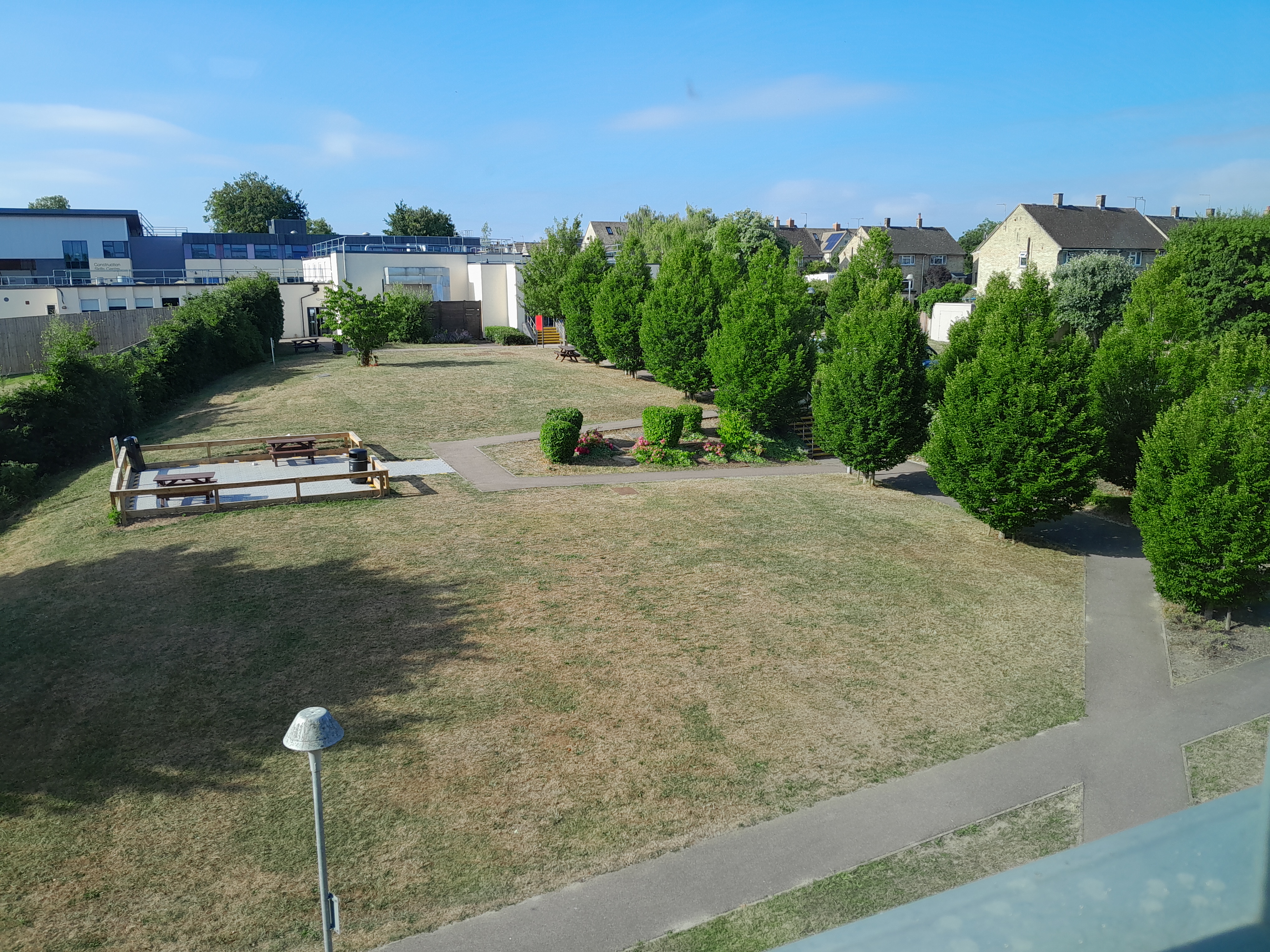 A view of the college from an upstairs window. There is a large grassy area, a fenced off smoking area, a tree-lined avenue and zig-zagging tarmac paths. There are three modern buildings in the background, one of which is labelled 'construction skills center'.